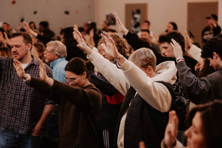 A crowd of people with raised hands at a gathering, some engaging in a moment of expression. The setting appears to be indoors, with muted colors in the background, and attendees displaying various styles of clothing.