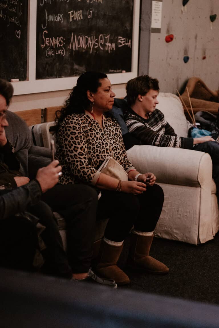 A woman with curly black hair sits on a couch wearing a leopard print top and holding a bag. Next to her, a young man in a striped sweater is seated, looking down at his phone. The interior has a chalkboard and climbing wall elements in the background.