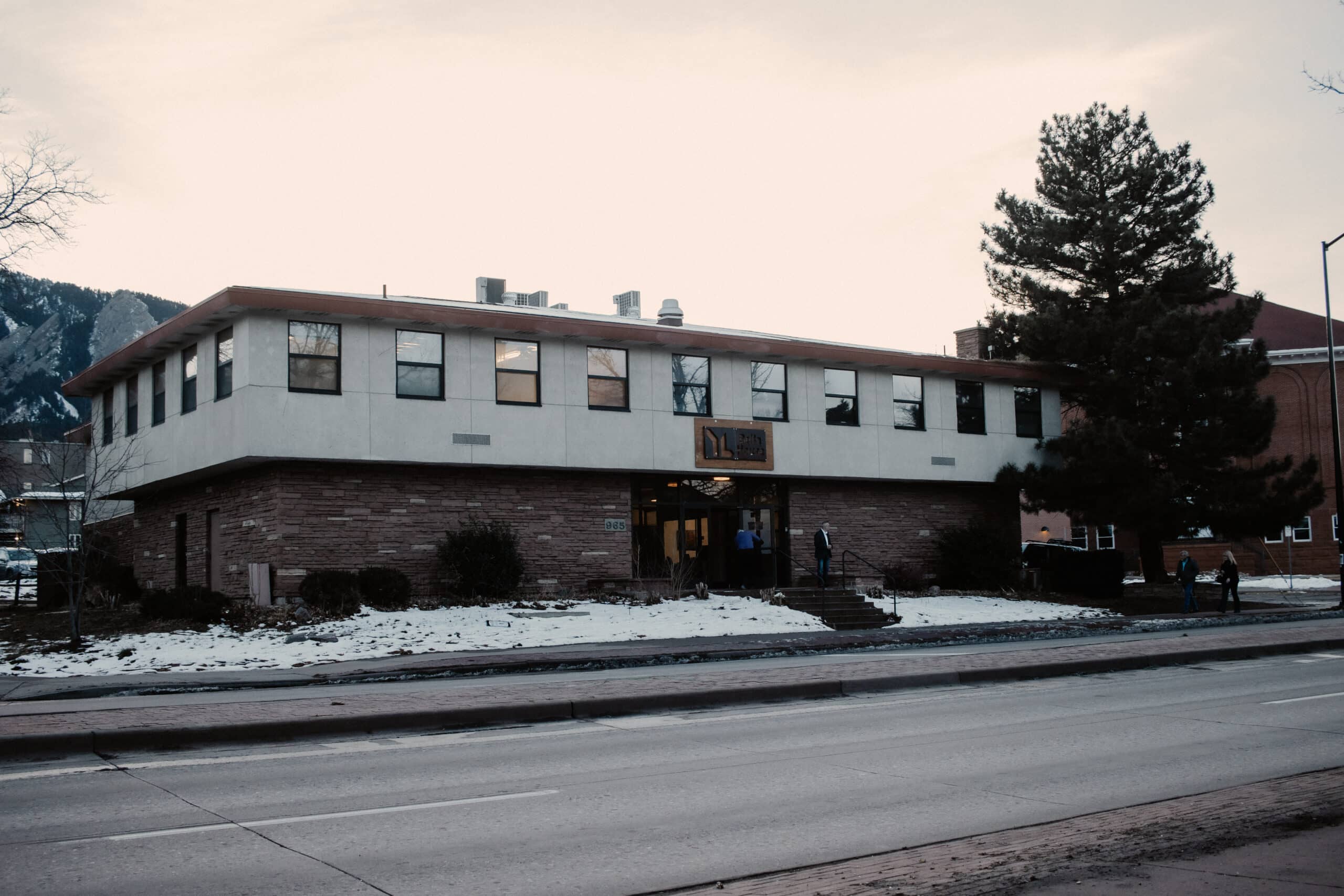 A two-story building with a stone and white exterior is viewed from the street. Large windows are visible on the upper floor. Snow covers the ground in front of the structure, and a few people are seen entering the building. Pine trees are nearby.