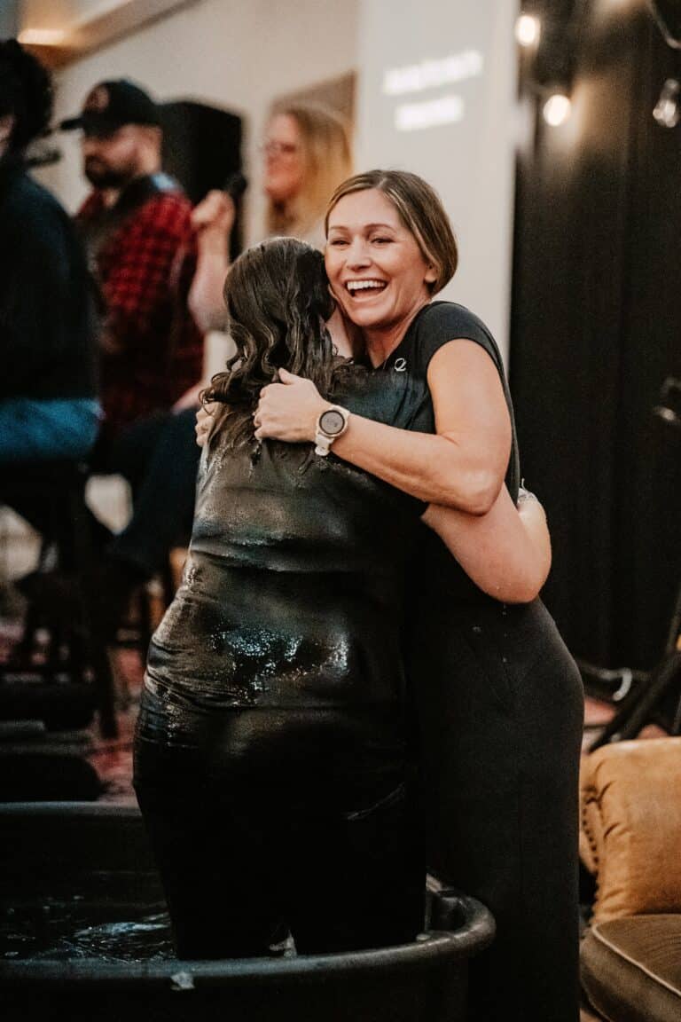 Two women embrace joyfully, one standing in a large tub and wet, while the other is outside the tub. They both smile and appear to be celebrating a significant moment. In the background, people are seated, including a musician on stage.