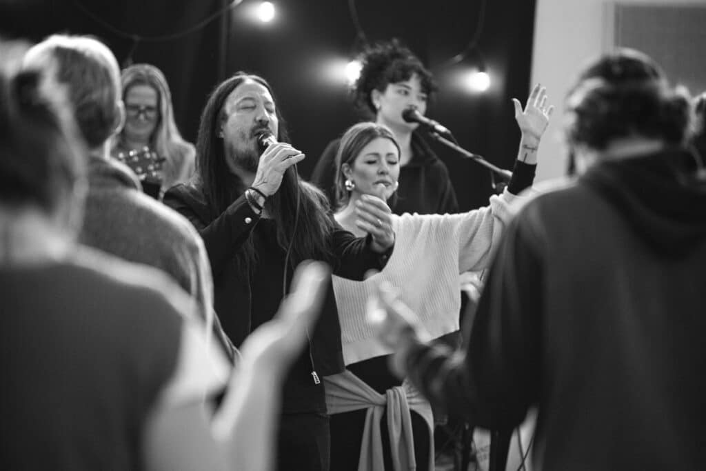 A monochrome image of a group of people singing in a collaborative setting. Two individuals are in the foreground: a man singing into a microphone and a woman with her hands raised. Others are engaged in the background, with instruments visible.
