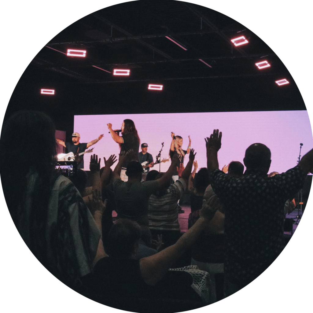 A group of people in a dimly lit room with their arms raised while participating in a live music performance. Two performers on stage are singing, with a large screen in the background displaying a soft pink light. The audience appears engaged.