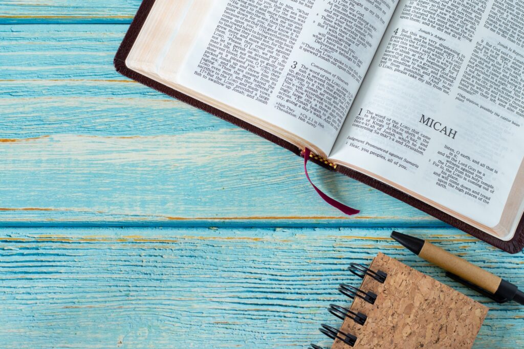 An open Bible on a blue wooden surface, with a focus on the page containing the word "MICAH." Beside the Bible is a small cork notebook and a black pen, arranged neatly on the table.