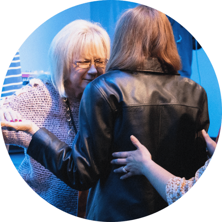 An elderly woman with short blonde hair and glasses appears to be engaging warmly with a woman in a black leather jacket, surrounded by others in a casual setting. The atmosphere suggests a moment of connection.