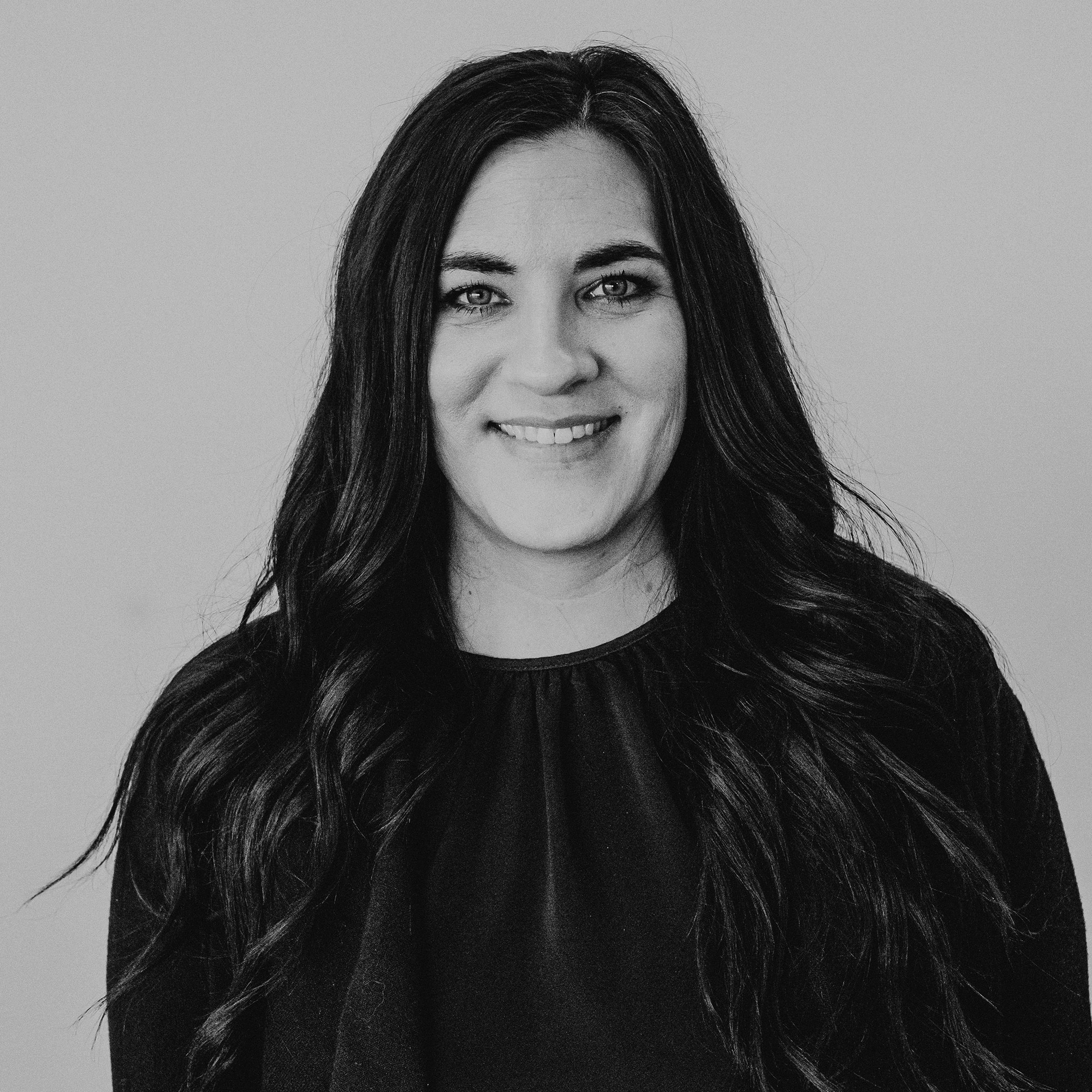 A woman with long, dark hair smiling at the camera. She is wearing a black top and has a neutral expression against a light gray background, highlighting her features and hairstyle.
