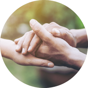 Two hands clasping gently, one hand positioned above the other, with soft fingers touching. The background is blurred greenery, creating a warm and intimate atmosphere. The hands represent connection and support.