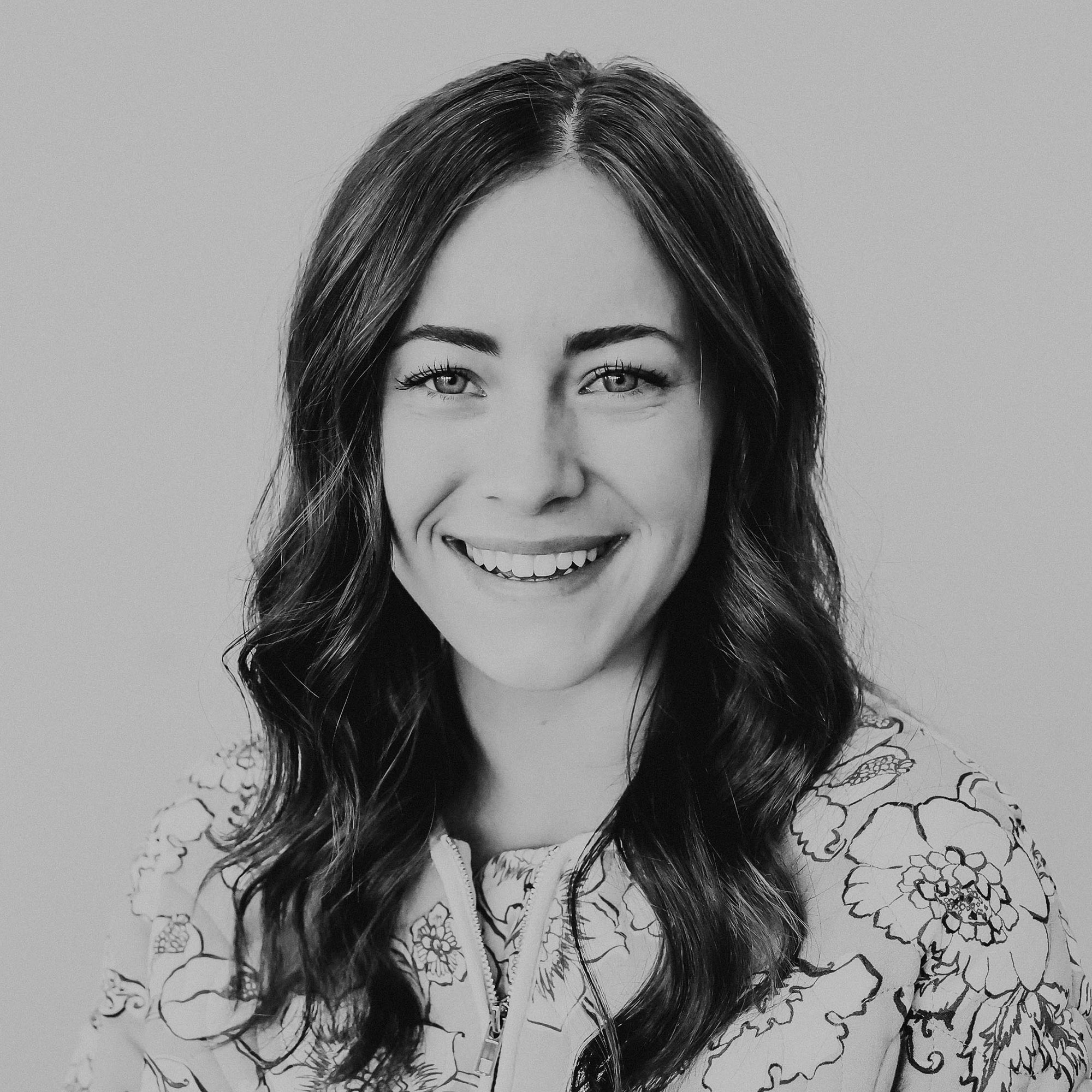 A woman smiling with long, wavy hair, wearing a floral-patterned top. The image is in black and white, showcasing her bright eyes and friendly expression against a light-colored background.