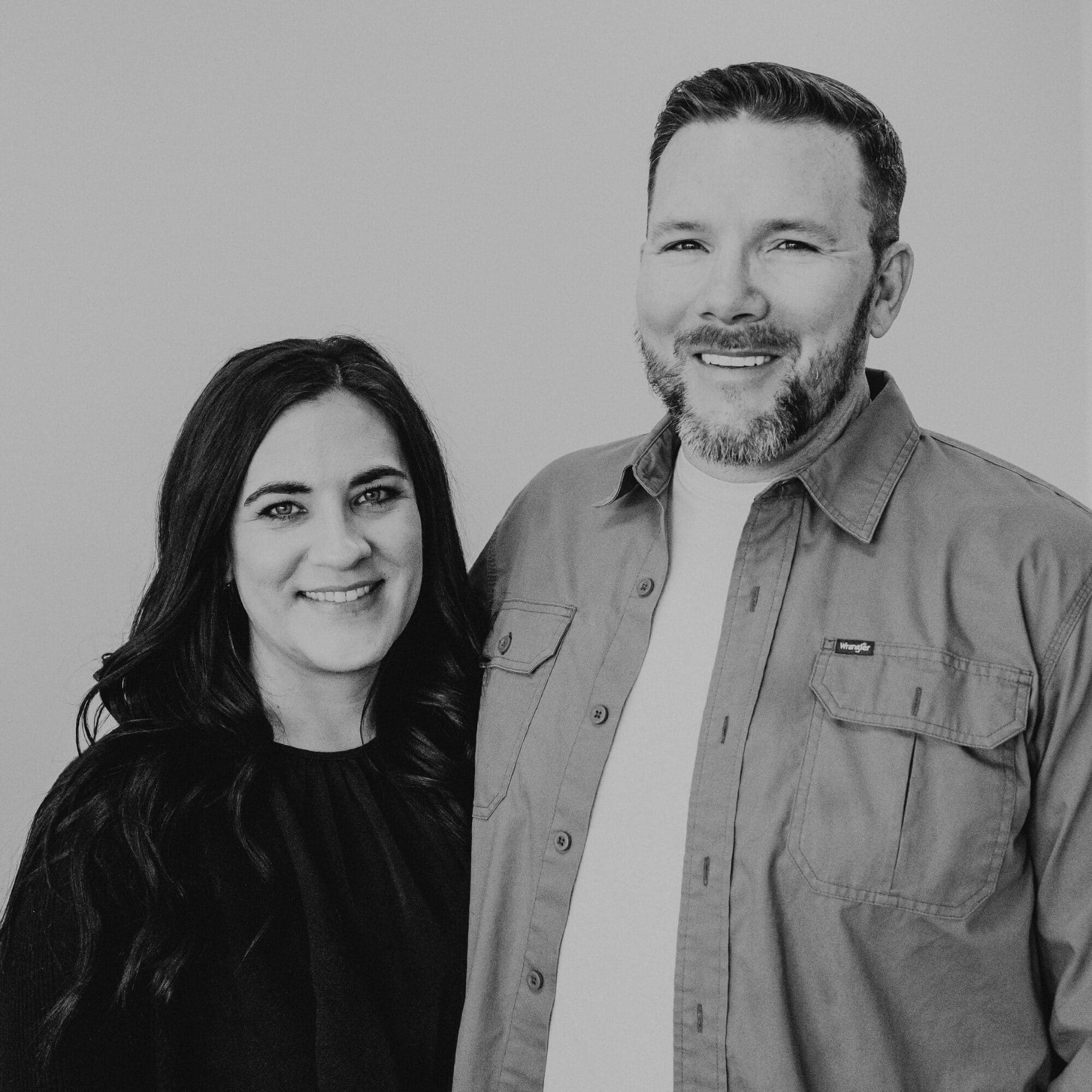 A woman with long dark hair and a man with short hair and a beard stand together, both smiling and looking at the camera. The background is neutral, and they are dressed casually, with the man wearing a button-up shirt over a plain t-shirt.