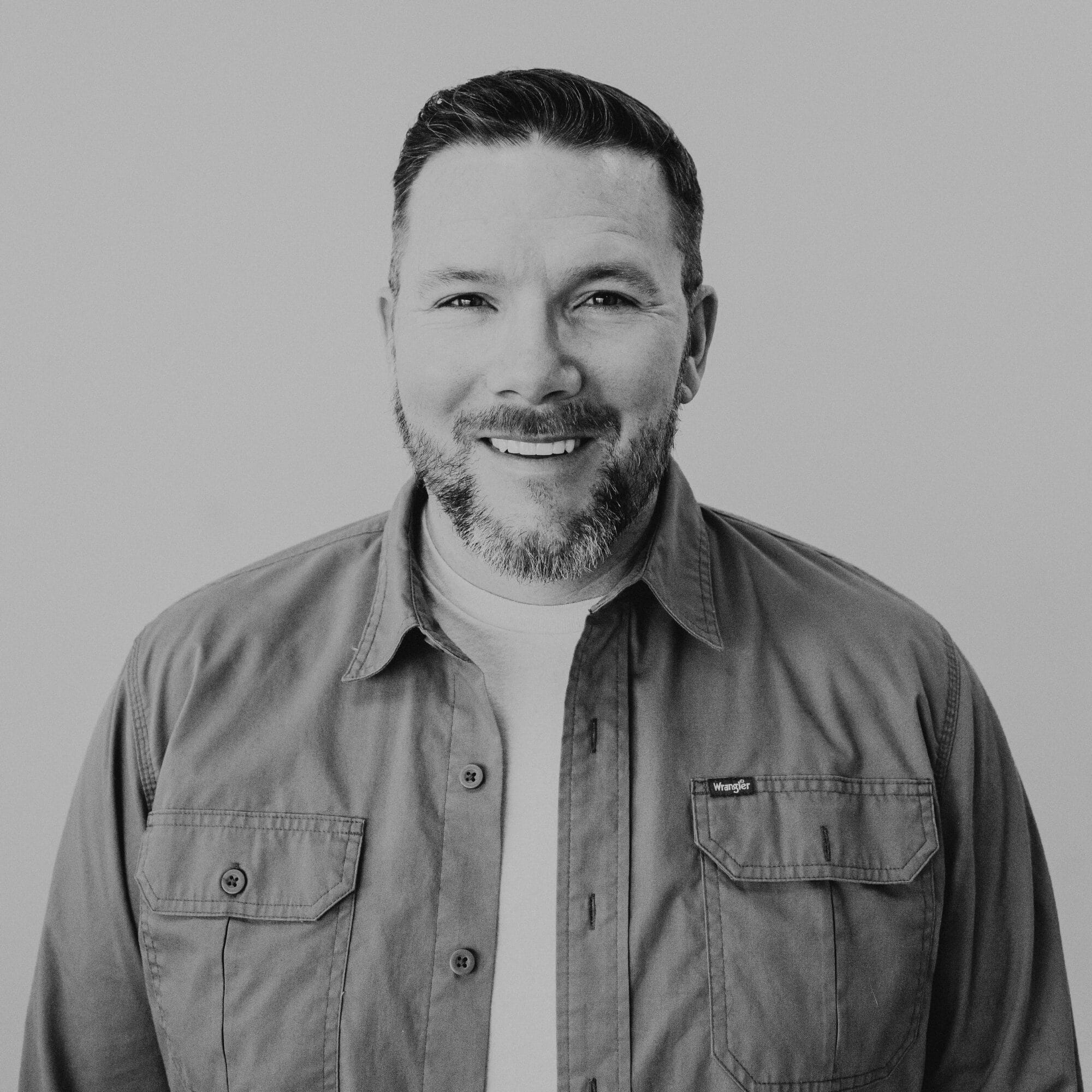 A smiling man with a short beard, wearing a light-colored shirt and a gray button-up jacket, stands in front of a plain light background. He has short, neatly styled hair and appears friendly and approachable.