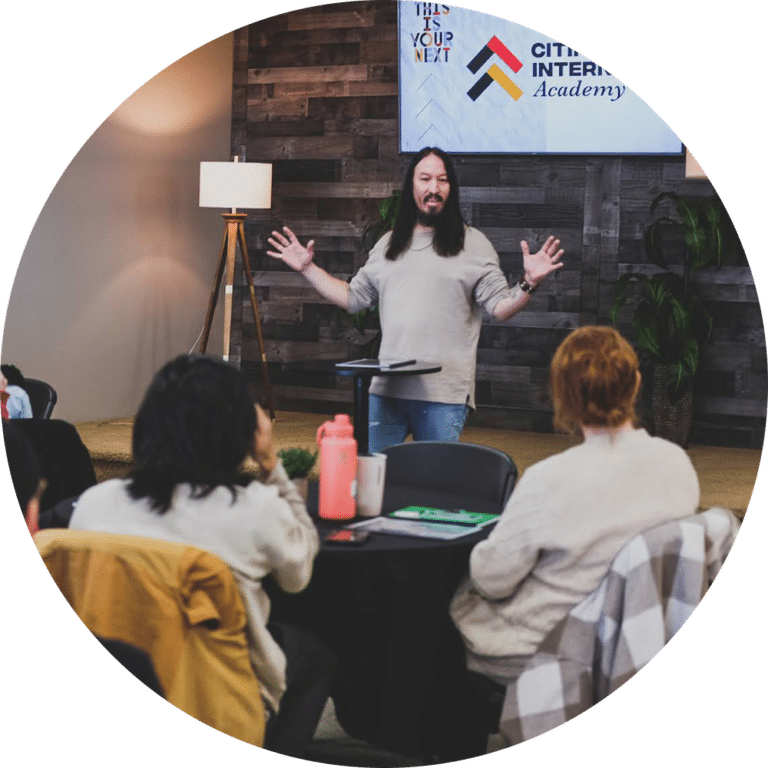 A speaker with long hair presents in front of an audience seated at tables. A large screen displays the text "Citizens International Academy" above the presenter. The setting features wooden walls and a bright lamp.