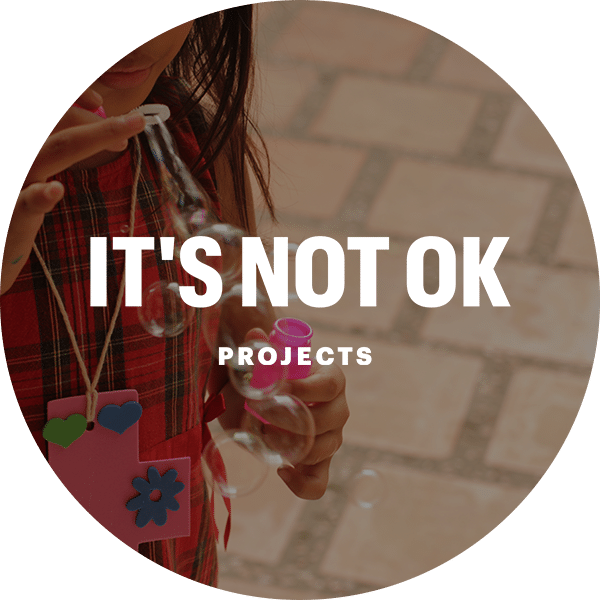 A person in a plaid shirt blowing bubbles with a bubble wand, holding a colorful necklace. The image features the text "IT'S NOT OK PROJECTS" prominently displayed in white font over a blurred background of stone pavement.