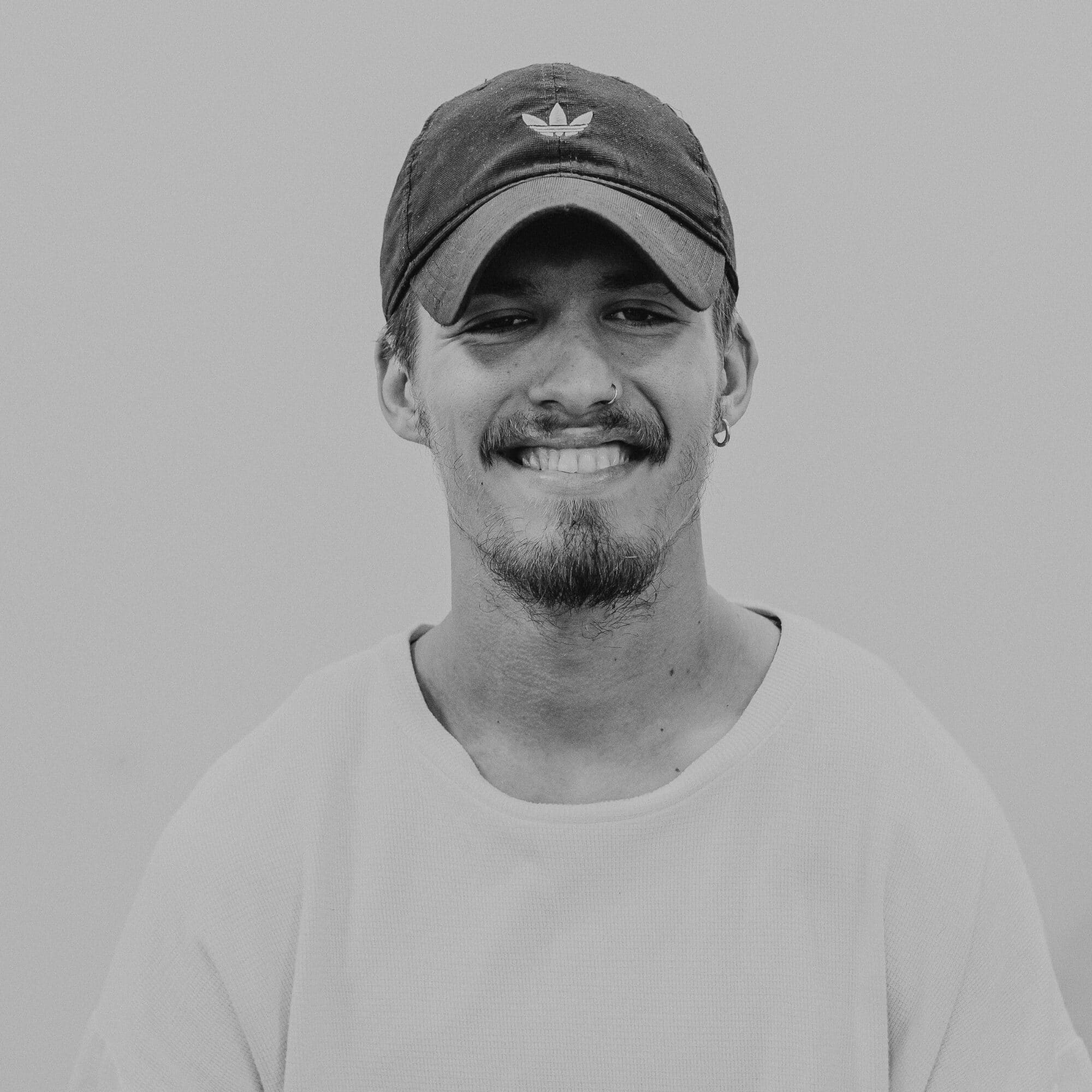 A young man wearing a black cap and a light-colored T-shirt. He has a beard and is smiling directly at the camera. The background is neutral and untextured, emphasizing the subject's facial features and expression.