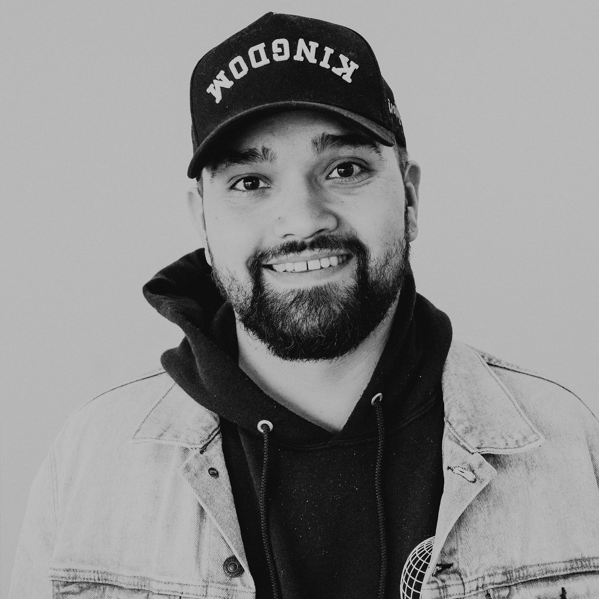 A young man wearing a black cap with the word "KINGDOM" on it, a dark hoodie, and a light denim jacket. He is smiling and standing against a neutral background, with short black hair and a beard.