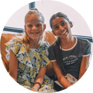 Two girls sitting on a couch, smiling at the camera. One girl has long blonde hair and is wearing a dress with lemon patterns. The other girl has long dark hair tied in a ponytail and is wearing a navy blue shirt with white text.
