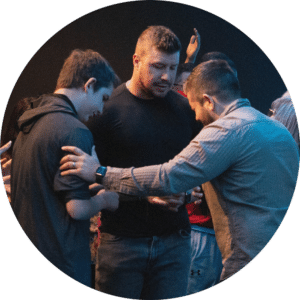 Two men and a teenage boy engage in a supportive gesture during a group gathering. The group is shown in a close-up setting with a dimly lit background. The men appear to be offering guidance or encouragement while standing close together.