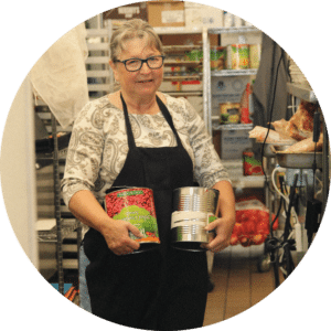 A woman wearing glasses and an apron is standing in a kitchen, holding two cans of food. She has a patterned shirt and appears to be in a food preparation area with shelves of ingredients visible in the background.