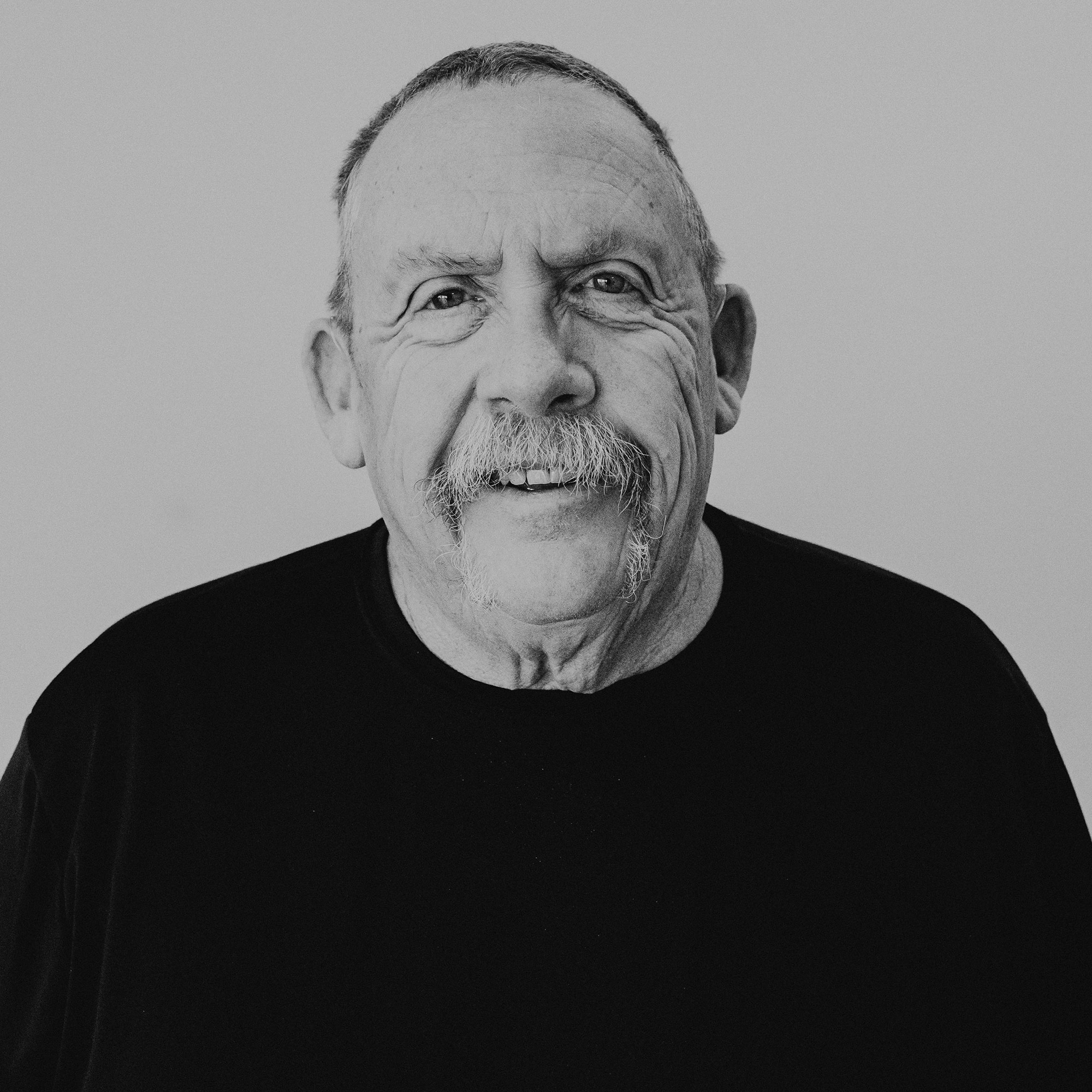 A senior man with a prominent mustache is smiling at the camera. He is wearing a black shirt against a light background. The image is in black and white, highlighting the man's features.
