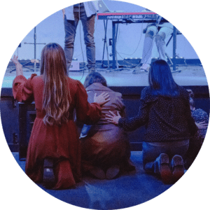 A group of three women kneeling in prayer or reflection at a stage, with their hands placed on each other's backs. The stage features a musician playing a keyboard in the background, illuminated by blue lighting.
