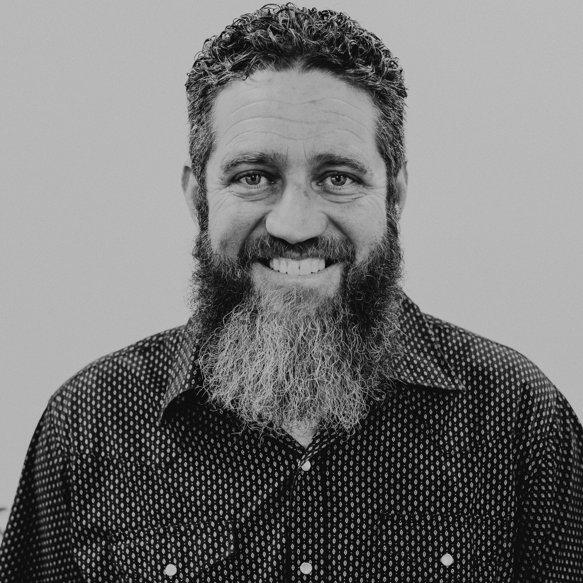 A smiling man with a full beard and curly hair is wearing a patterned black shirt. He is posing against a neutral gray background, looking directly at the camera. His expression is friendly and approachable.