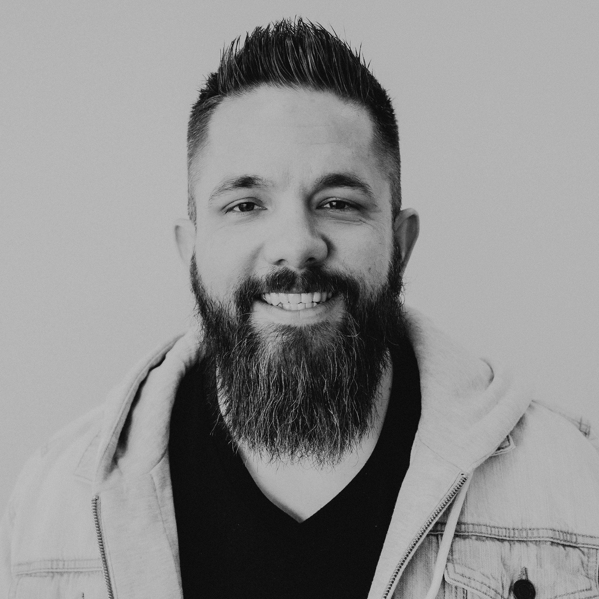 A man with a full beard and short hair smiles at the camera. He is wearing a black V-neck shirt and a light-colored denim jacket. The background is neutral, enhancing the focus on his facial features.
