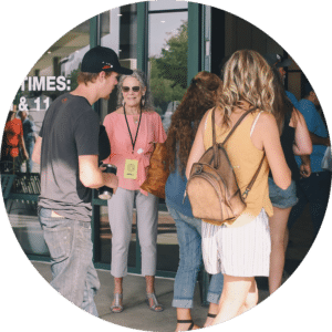 A group of people entering a building. In the foreground, a woman with glasses and curly hair greets them, holding a sign. A young man in a black shirt and a girl with long, wavy hair are visible, along with others in casual clothing.