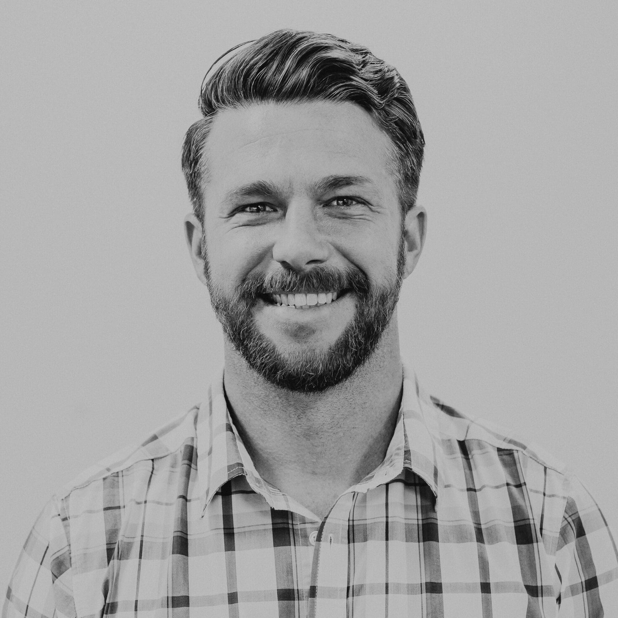 A smiling man with a beard and short hair posing for a portrait. He is wearing a plaid shirt and is set against a plain background, creating a clean and professional appearance.