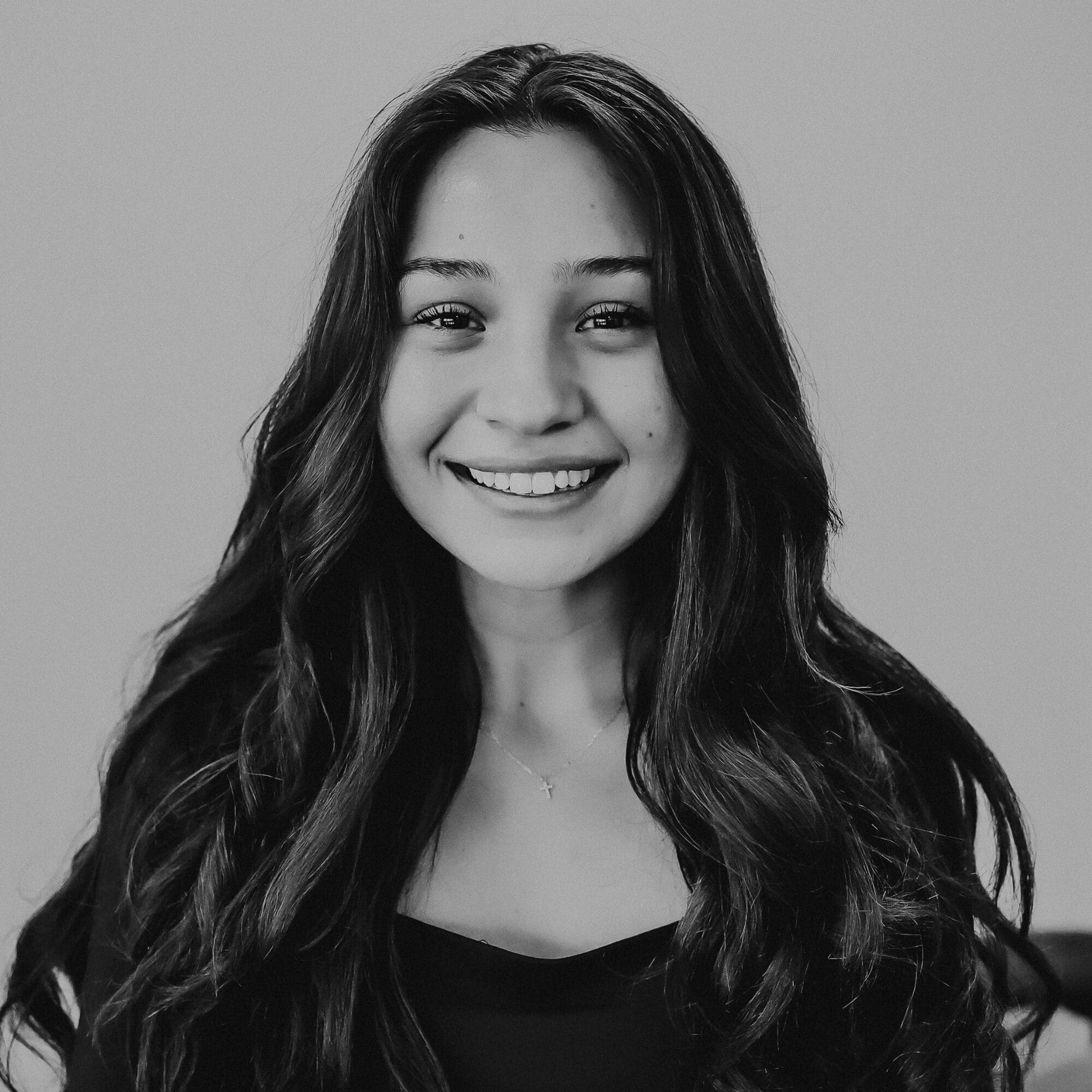 A smiling young woman with long, wavy dark hair and a subtle necklace is standing against a light background. She is wearing a black top and appears happy, with her eyes bright and welcoming.