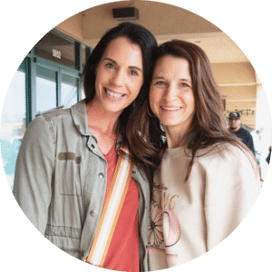 Two women standing closely together and smiling at the camera. The woman on the left has dark hair and is wearing a gray jacket over a red top. The woman on the right has light brown hair and is dressed in a light sweater. A building entrance is visible in the background.
