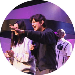 A young man passionately singing into a microphone on stage, with a woman beside him holding her hands together. Another man is singing in the background. The setting features colorful lighting and modern stage design.
