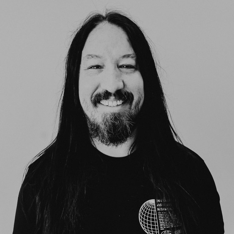 A man with long black hair and a beard smiles at the camera. He is wearing a black t-shirt with a graphic design. The background is neutral, highlighting his facial features.