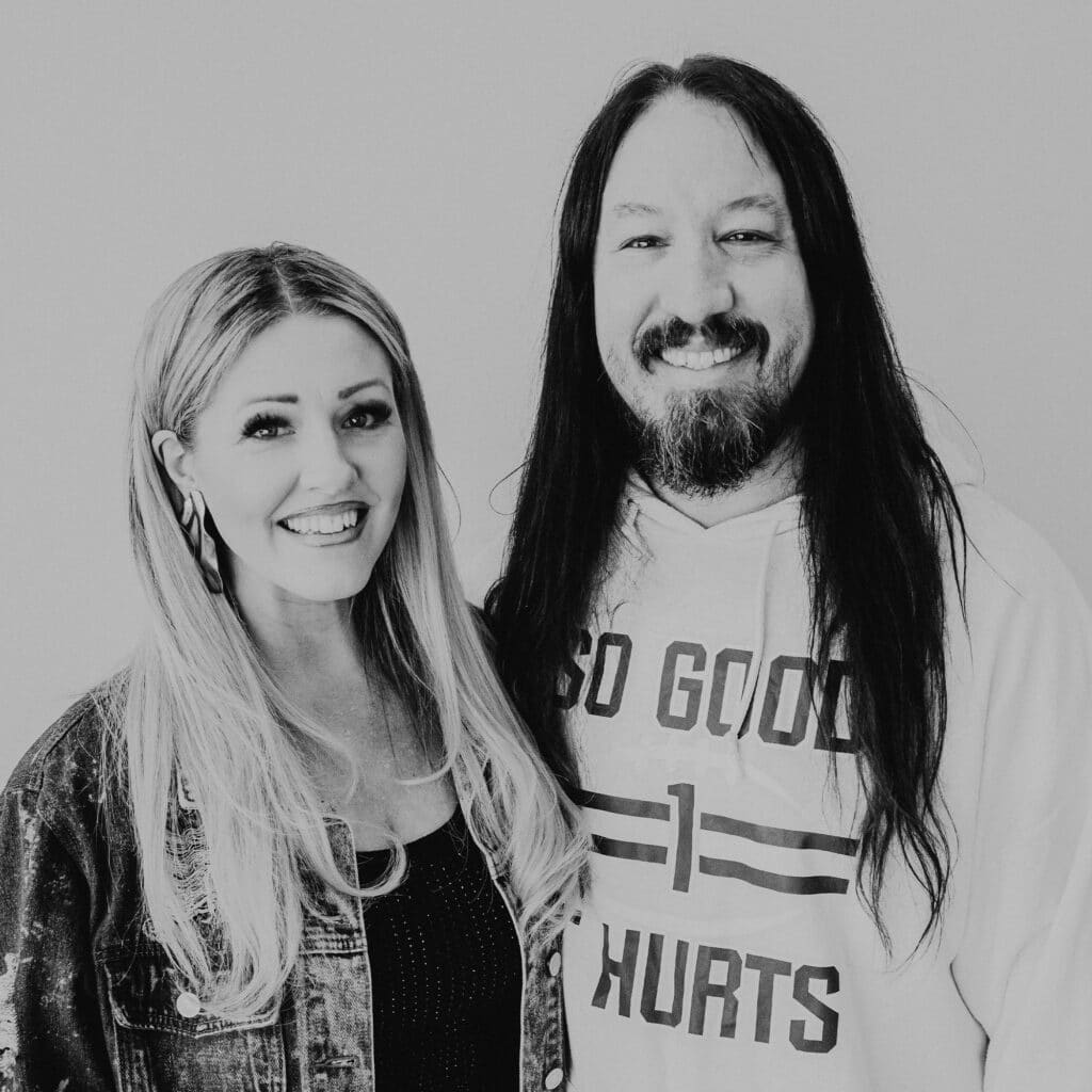 A woman with long blonde hair wearing a denim jacket and a black top stands next to a man with long black hair dressed in a white hoodie with text. They are both smiling against a light background.