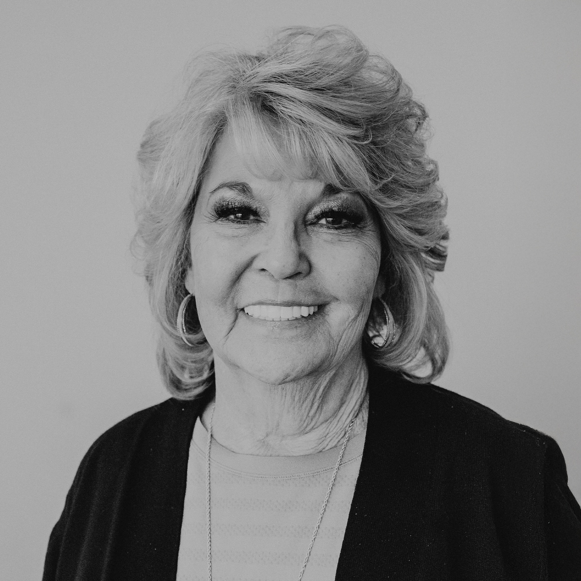 A smiling older woman with shoulder-length, wavy hair wearing a black cardigan over a light-colored top. She has a simple necklace and is positioned against a neutral backdrop.