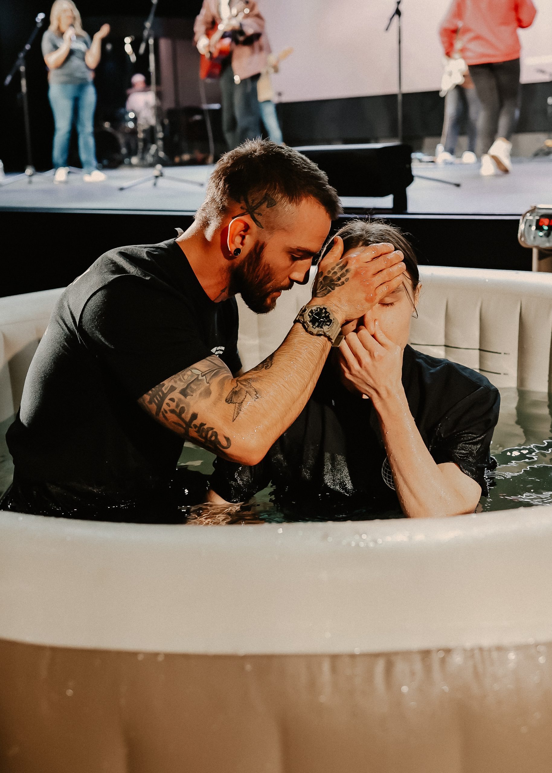 A man in a black shirt holds another man’s head under water in a circular inflatable baptismal pool. The scene shows people in the background, while the venue appears to have a stage with a band.