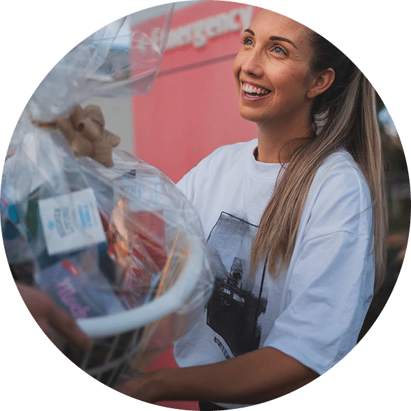A woman with long hair and a white t-shirt smiles as she receives a gift basket. The basket is wrapped in clear plastic and contains various items. A colorful backdrop with a faint logo can be seen behind her, contributing to an outdoor setting.