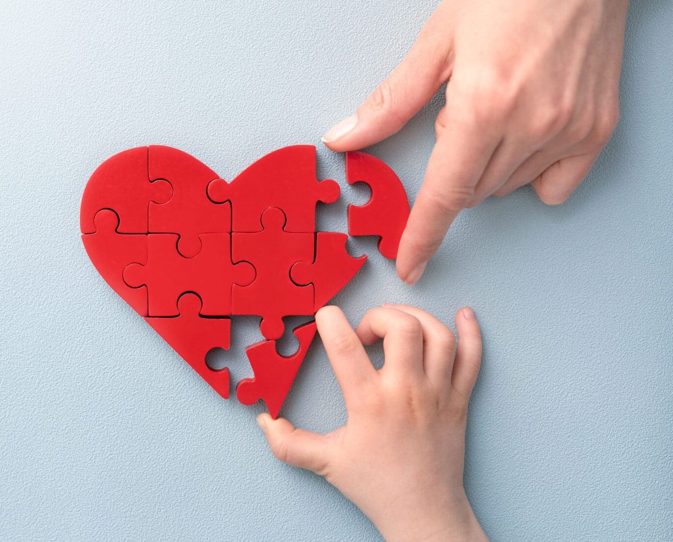 A puzzle shaped like a heart, made of red pieces, is being assembled. An adult’s hand is placing the final piece while a child's hand reaches out to help. The background is light blue, emphasizing the heart shape.