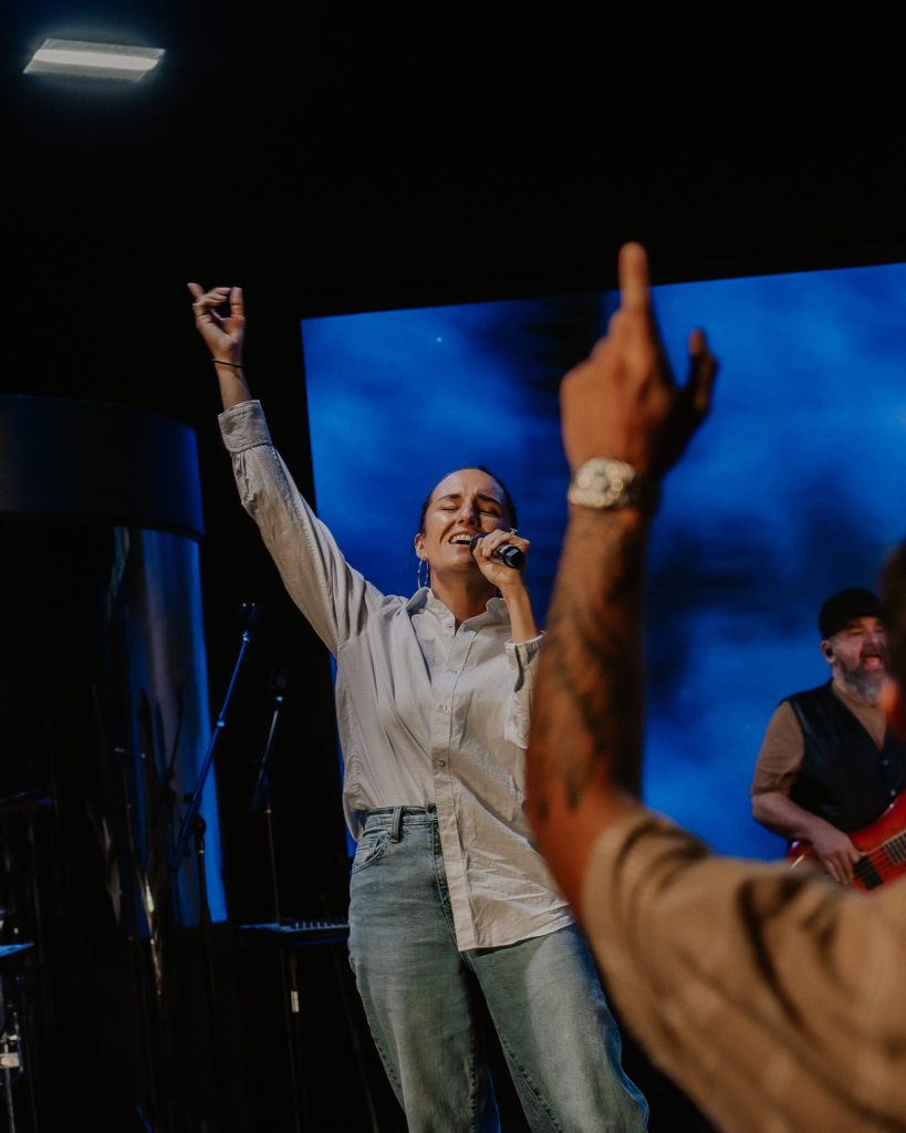 A female singer stands on stage, holding a microphone and raising one arm while performing. She wears a light blue shirt and jeans. In the background, a musician plays a bass guitar, and colorful lights create a vibrant atmosphere.