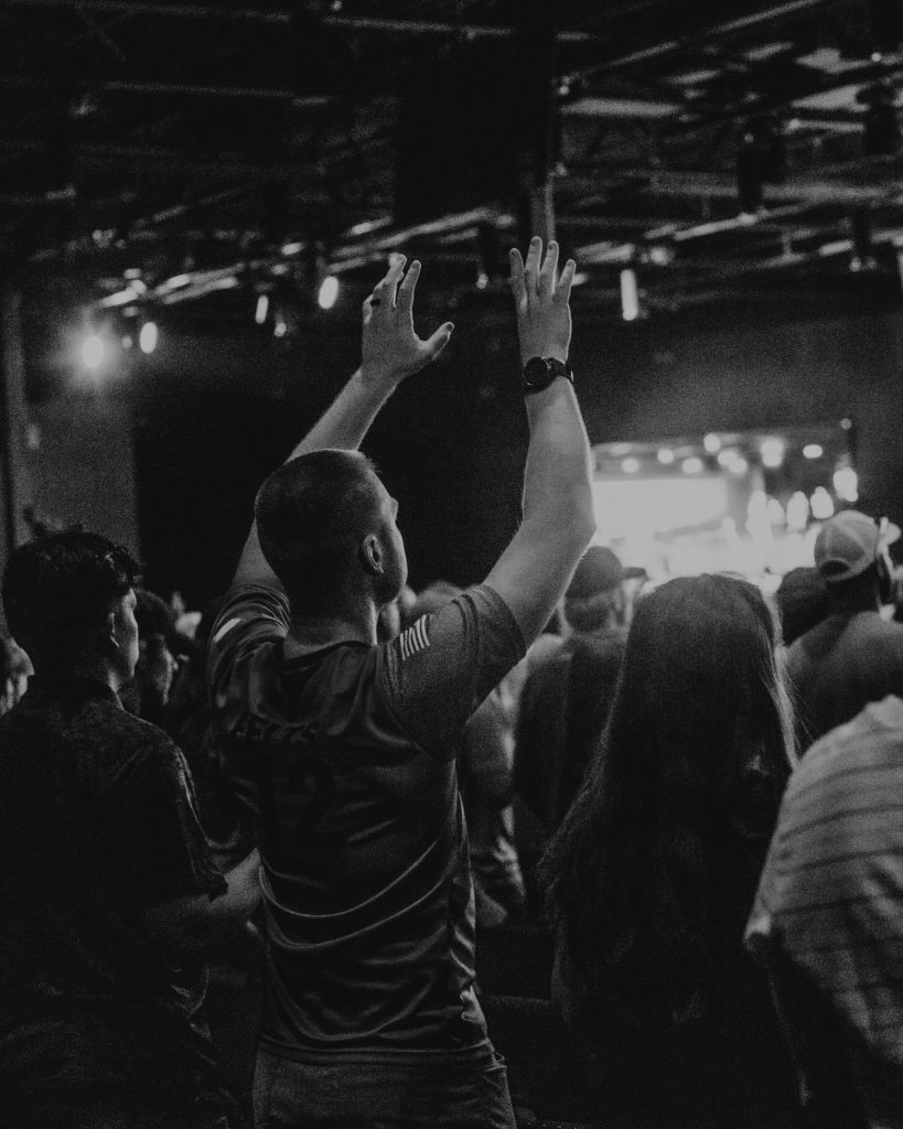 A man with short hair raises his hands in a crowd during a performance at an indoor venue. The audience is engaged, with some people facing the stage, which is illuminated with bright lights in the background.