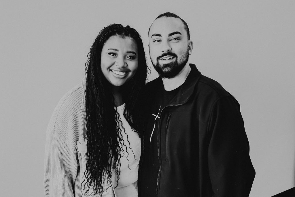 A black and white photo of a woman with long curly hair and a man with a beard, both smiling and standing close together. The background is light, creating a neutral atmosphere.