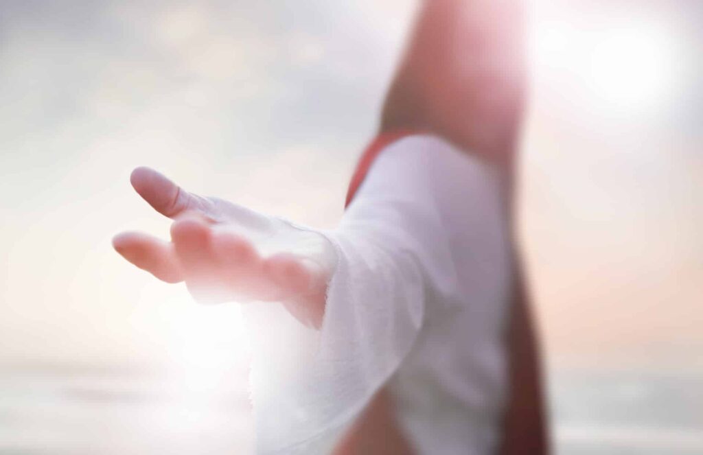 A figure in a white robe extends a hand forward, with a blurred ocean and soft sunlight in the background. The lighting creates a glowing effect around the figure, enhancing the serene atmosphere of the scene.