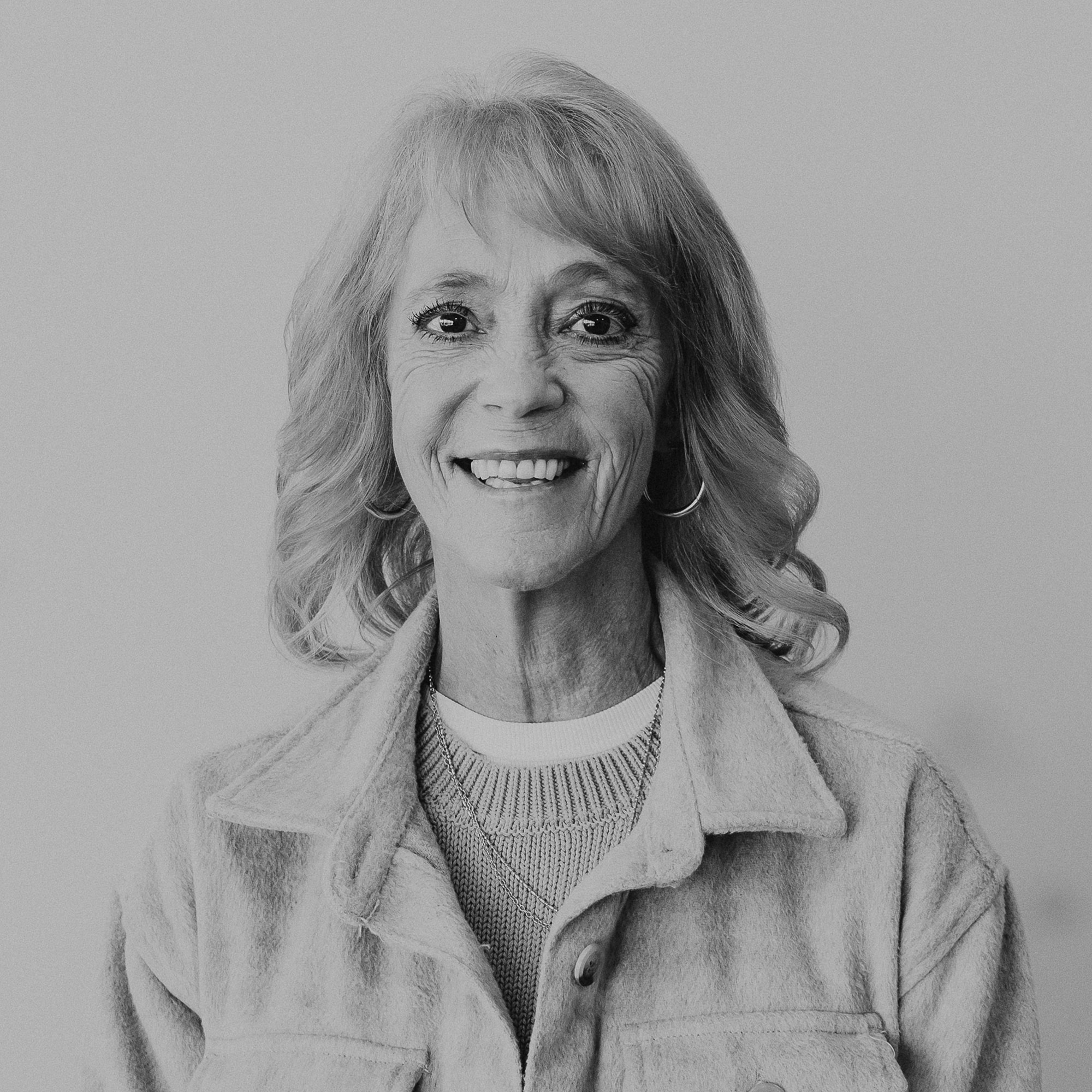 A woman with shoulder-length blonde hair and a warm smile, wearing a light-colored jacket over a sweater. The background is plain and light-colored, emphasizing her expression.