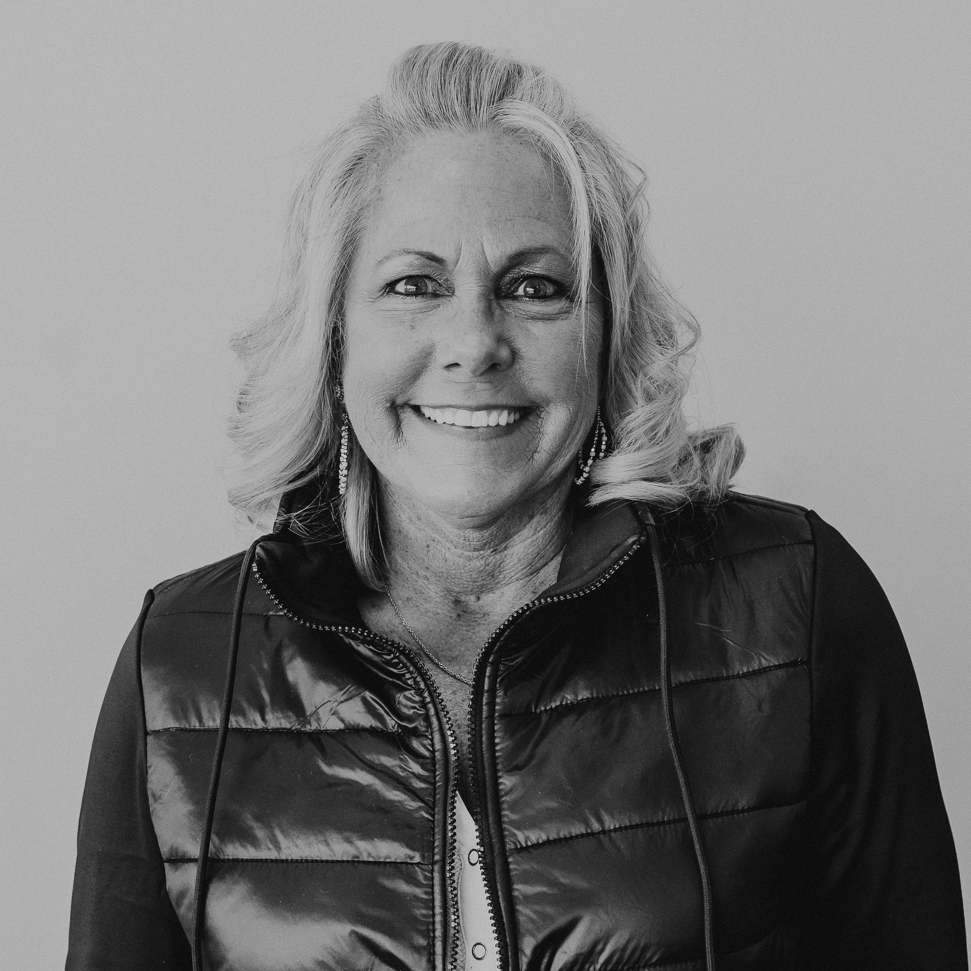 A woman with shoulder-length blonde hair smiling, wearing a black zip-up vest over a white shirt. The background is a neutral light color, giving focus to her expression and attire. She appears relaxed and approachable.