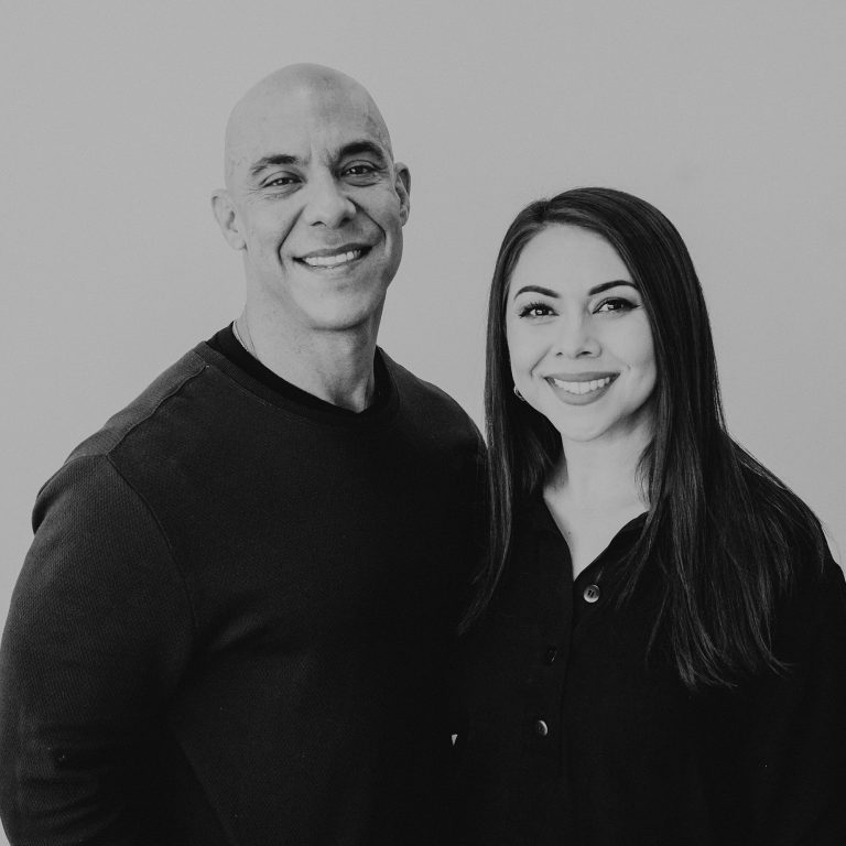 A man and a woman smiling at the camera, standing closely together against a plain gray background. The man has a bald head and is wearing a black long-sleeve shirt. The woman has long dark hair and is dressed in a black button-up top.