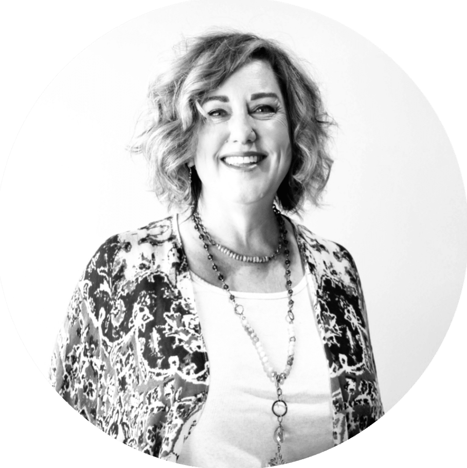 A woman with curly hair smiles at the camera, wearing a patterned shawl over a white shirt. She has multiple necklaces and a neutral background. The image is in black and white with a circular crop.