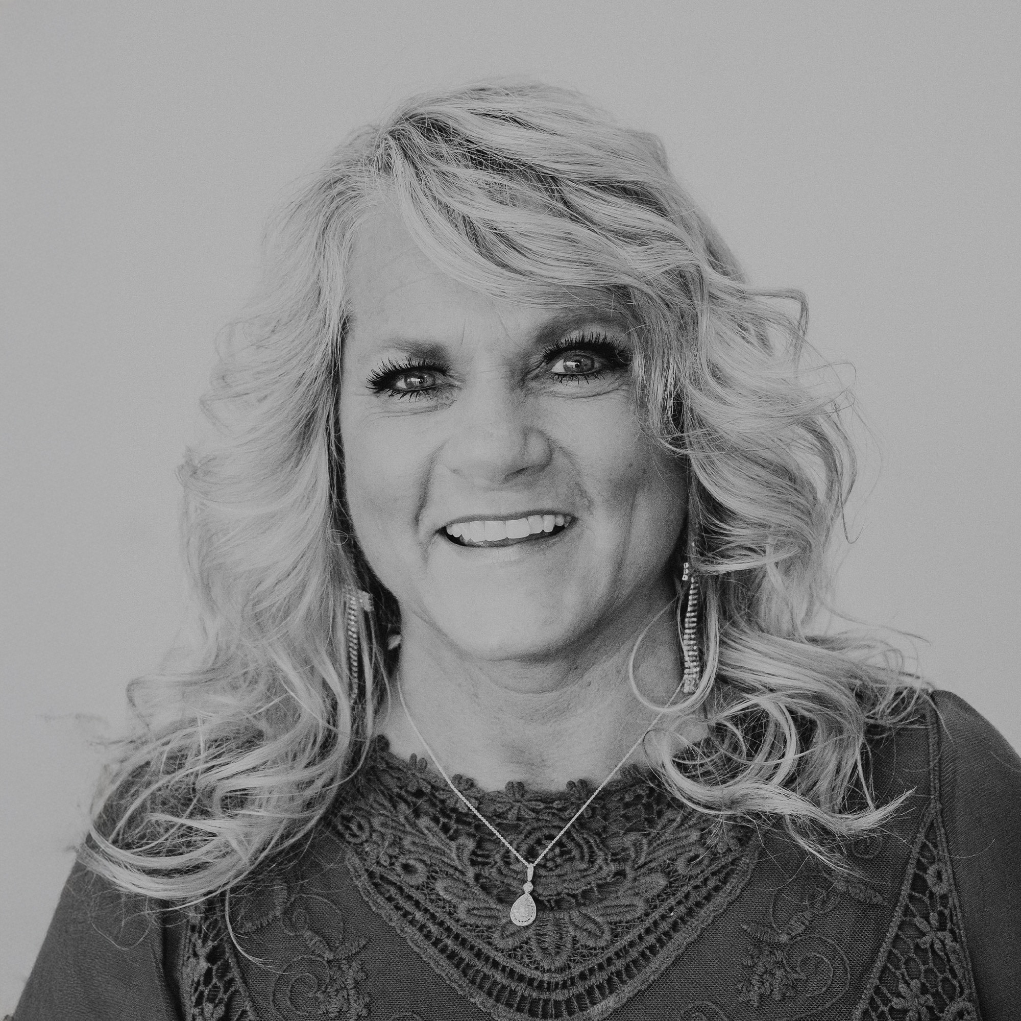 A middle-aged woman with long, curly blonde hair poses for a portrait. She is wearing large earrings and a necklace, with a smile on her face. The background is plain, creating a focus on her features.