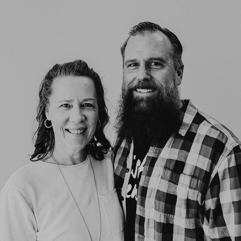 A woman with shoulder-length hair wearing a light-colored top and a man with a beard wearing a plaid shirt stand side by side, smiling at the camera. The background is a plain light color, highlighting their faces.