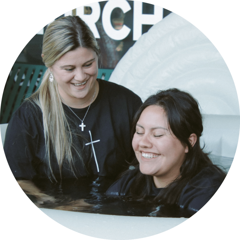 Two women in black shirts are standing in a baptismal pool. One woman, positioned behind, smiles while gently holding the other woman's shoulders, who appears relaxed and is smiling. The background includes elements that suggest a religious setting.